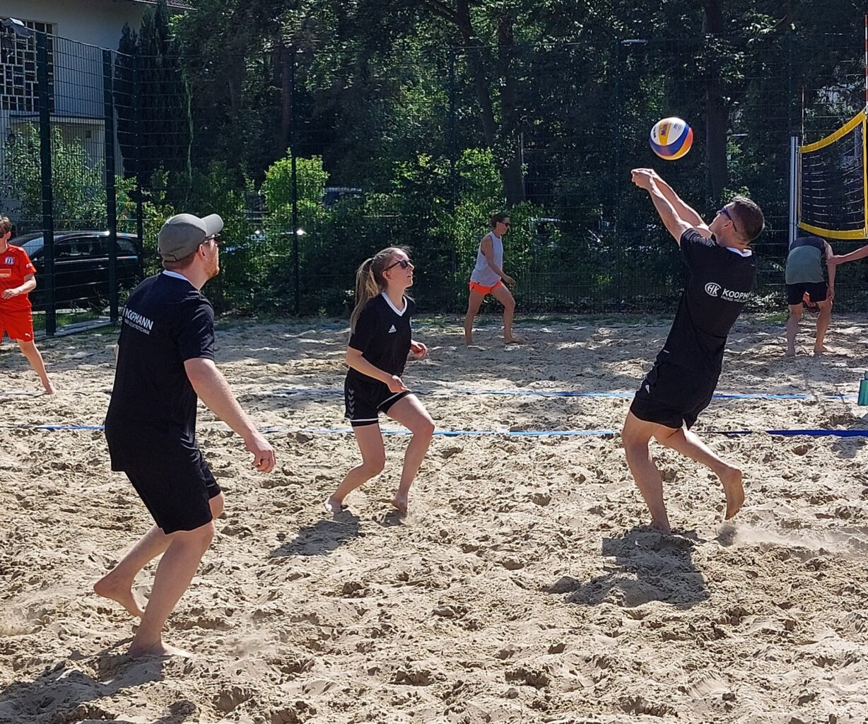Im Vordergrund spielen drei Personen in schwarzer kurzer Kleidung Volleyball auf einem Sandplatz und im Hintergrund spielen weitere Menschen Volleyball.
